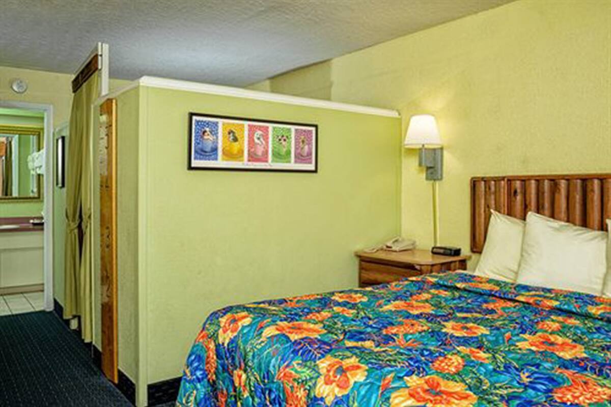 Une chambre d’hôtel avec un lit, une table de chevet, une lampe, un tableau encadré au mur et une porte.