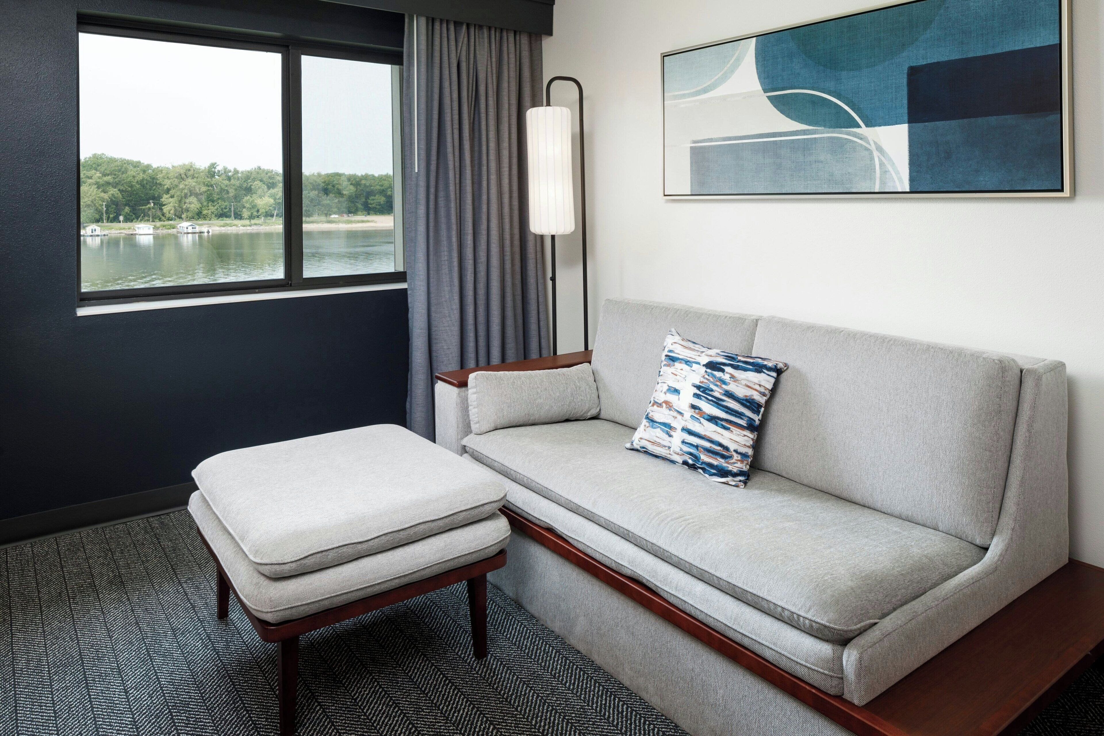 A modern hotel room with a grey sofa, a matching ottoman, a floor lamp, and a large window offering a view of a lake and trees.