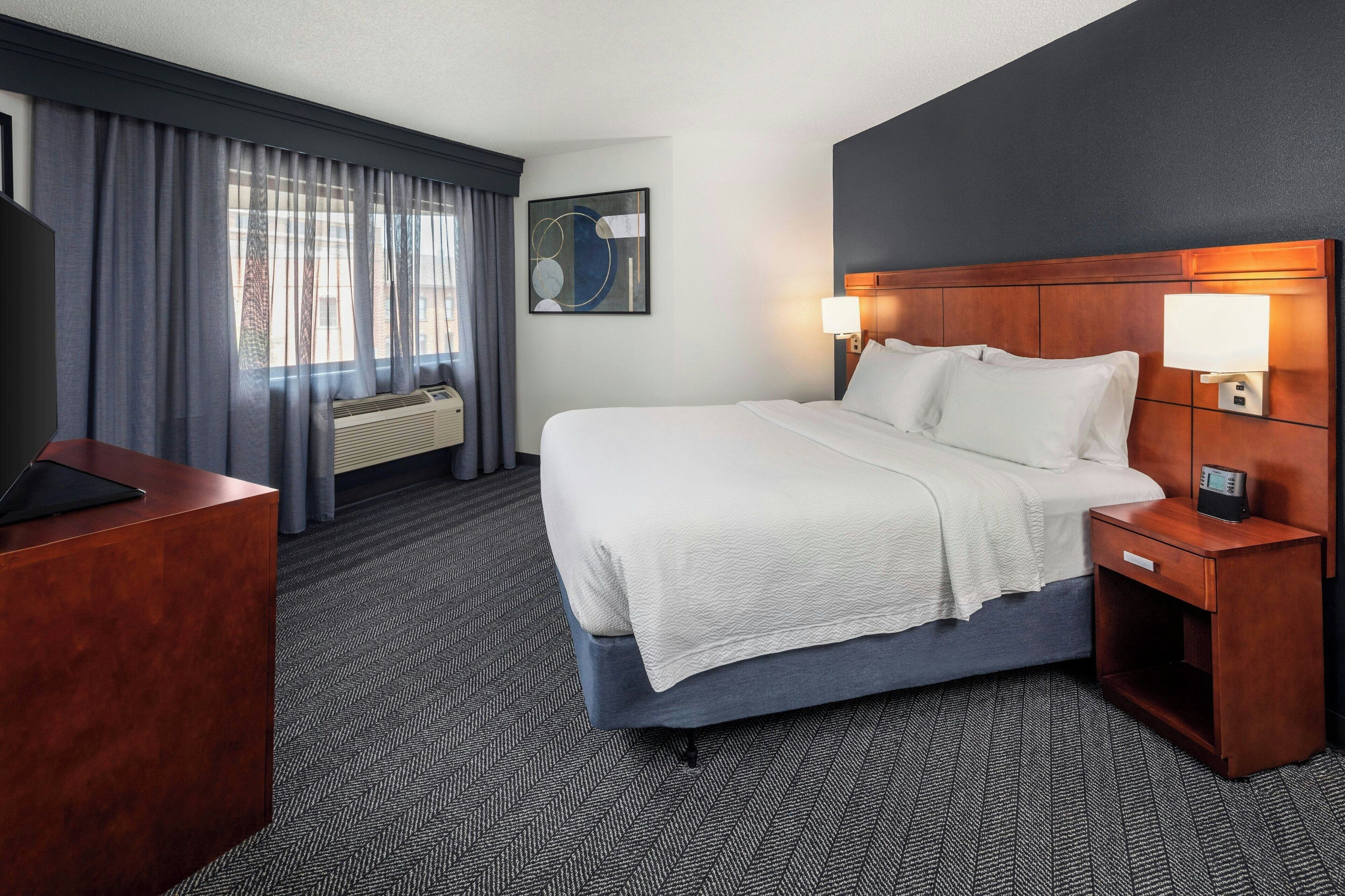 A hotel room with a large bed, a desk, a chair, a TV, and a window with curtains.
