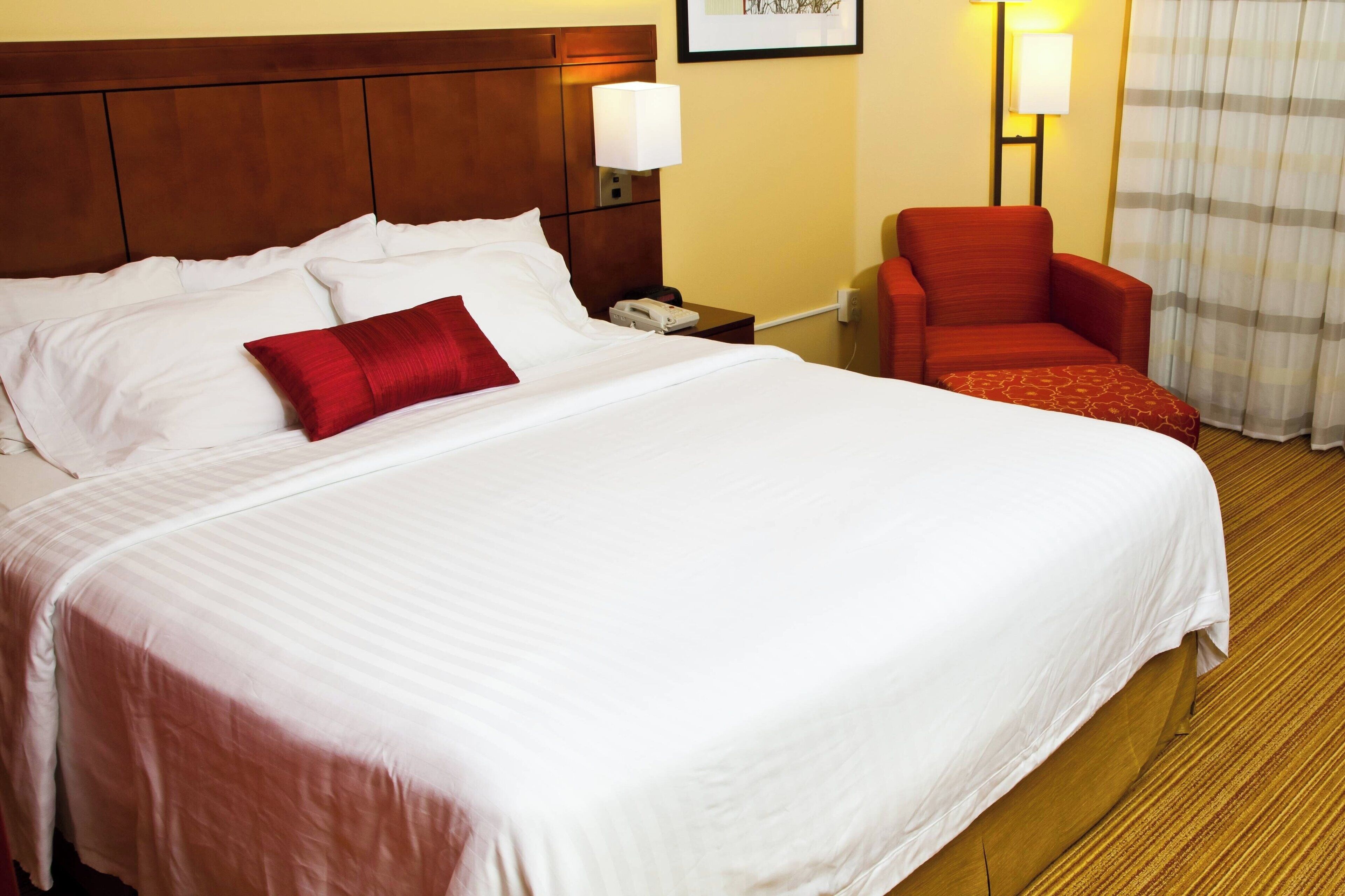 Habitación de hotel con una cama grande, una mesita de noche con lámpara, una silla roja y una ventana con cortinas.