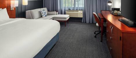 A hotel room with a large bed, a desk, a chair, a sofa, a TV, and a window with curtains.