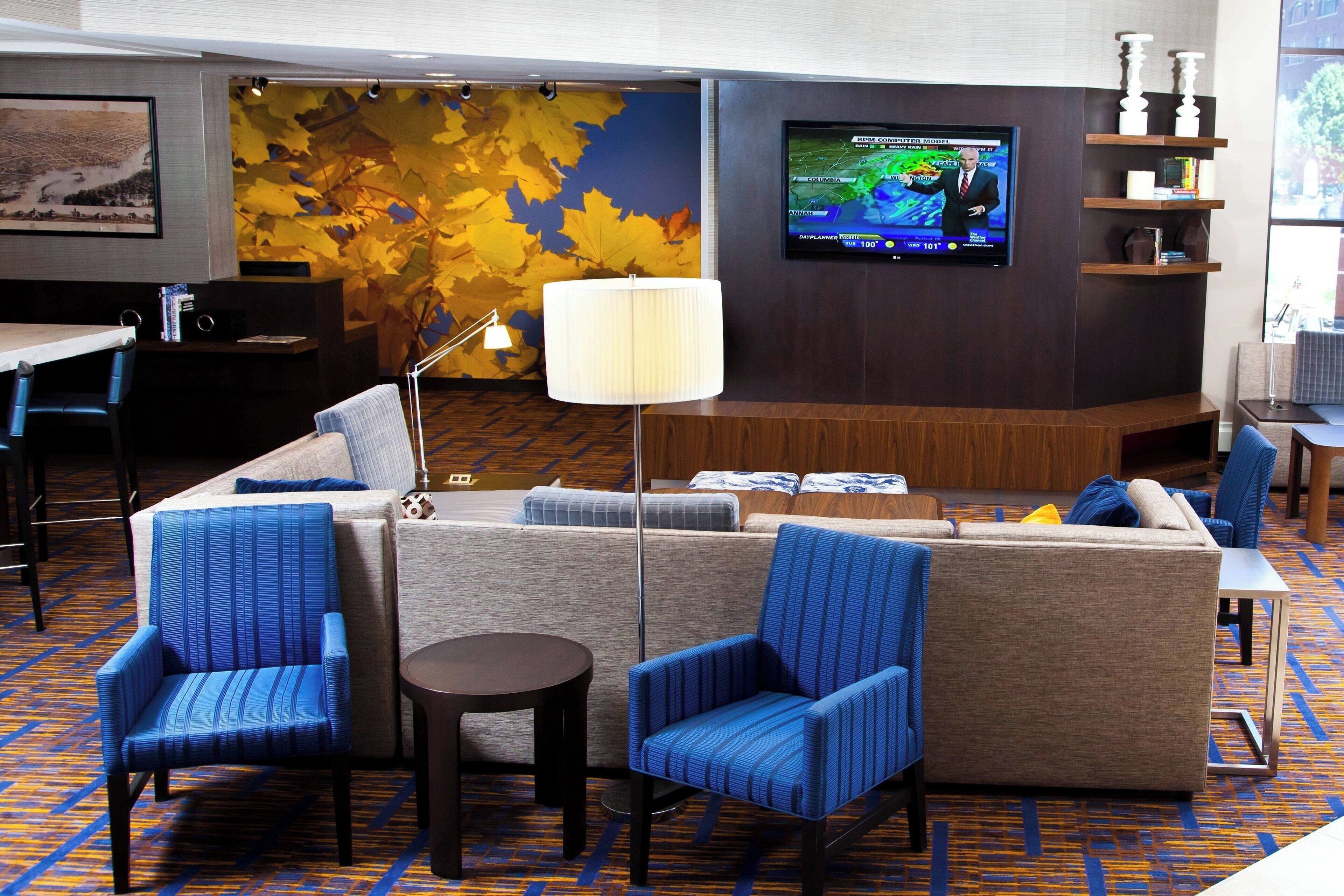 A hotel lobby with a large TV screen, seating area with blue chairs, a sofa, and a patterned carpet.
