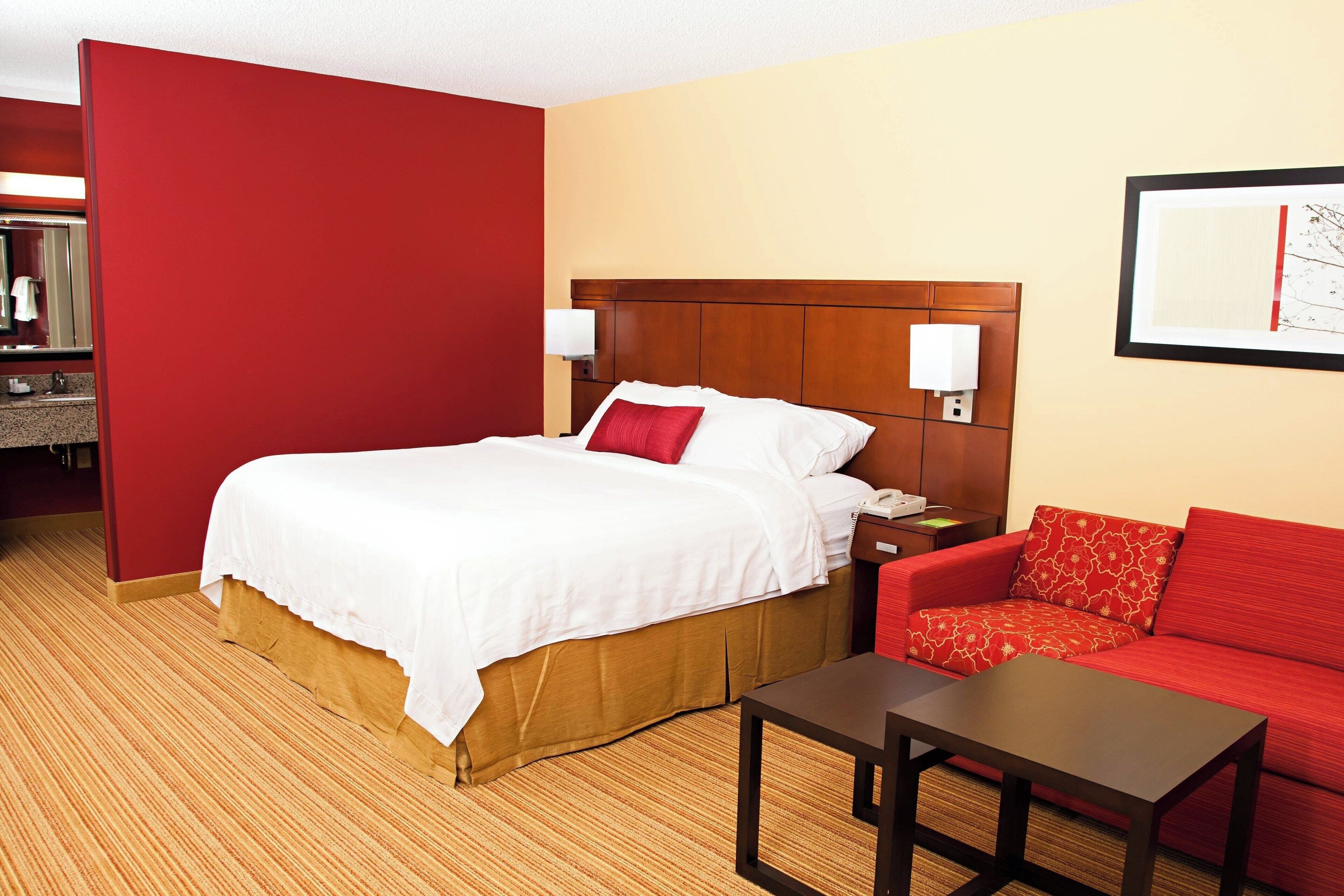 A hotel room with a bed, a red sofa, a small table, and a wall-mounted lamp.