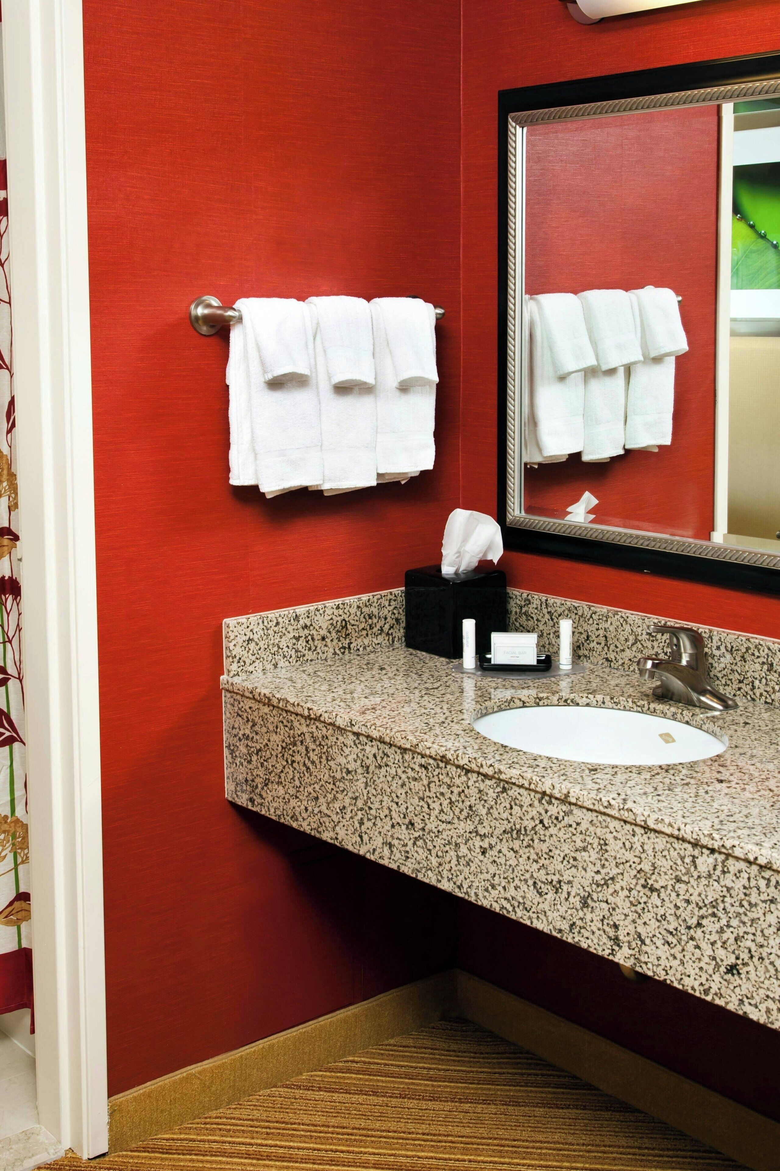 Un baño con una pared roja, un lavabo de granito, un espejo y toallas blancas colgadas en un perchero.