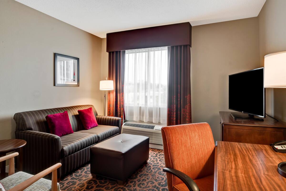 Habitación de hotel con sofá, otoman, sillón, mesa, televisión y ventana con cortinas.