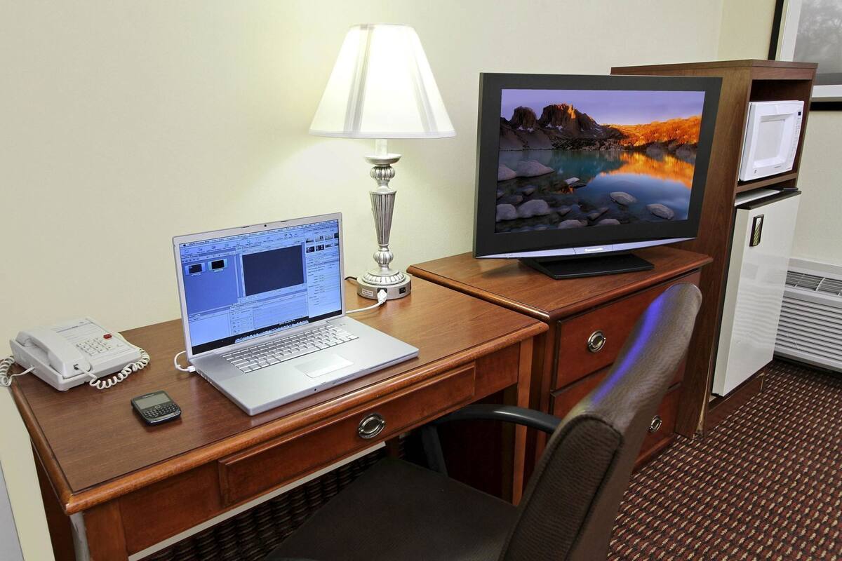 A hotel room with a wooden desk, a laptop, a desktop monitor, a telephone, a lamp, and a refrigerator.
