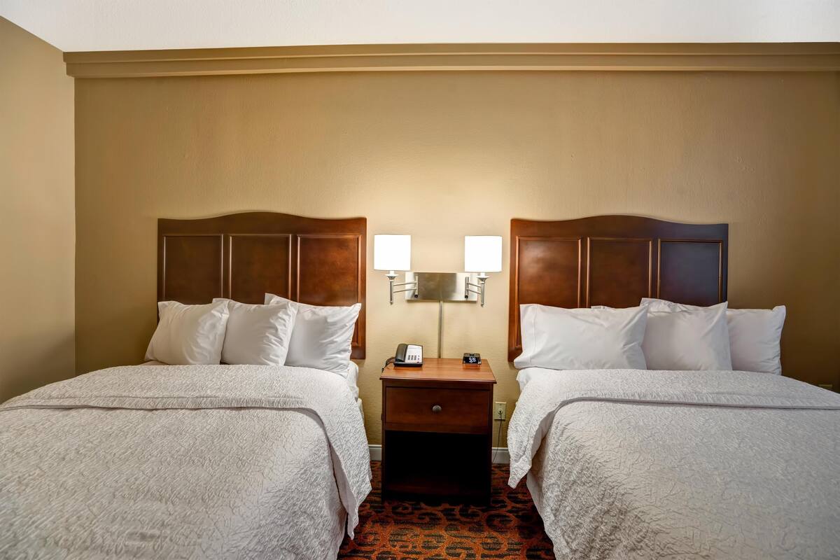 Una habitación de hotel con dos camas, una mesita de noche con lámpara y un reloj.