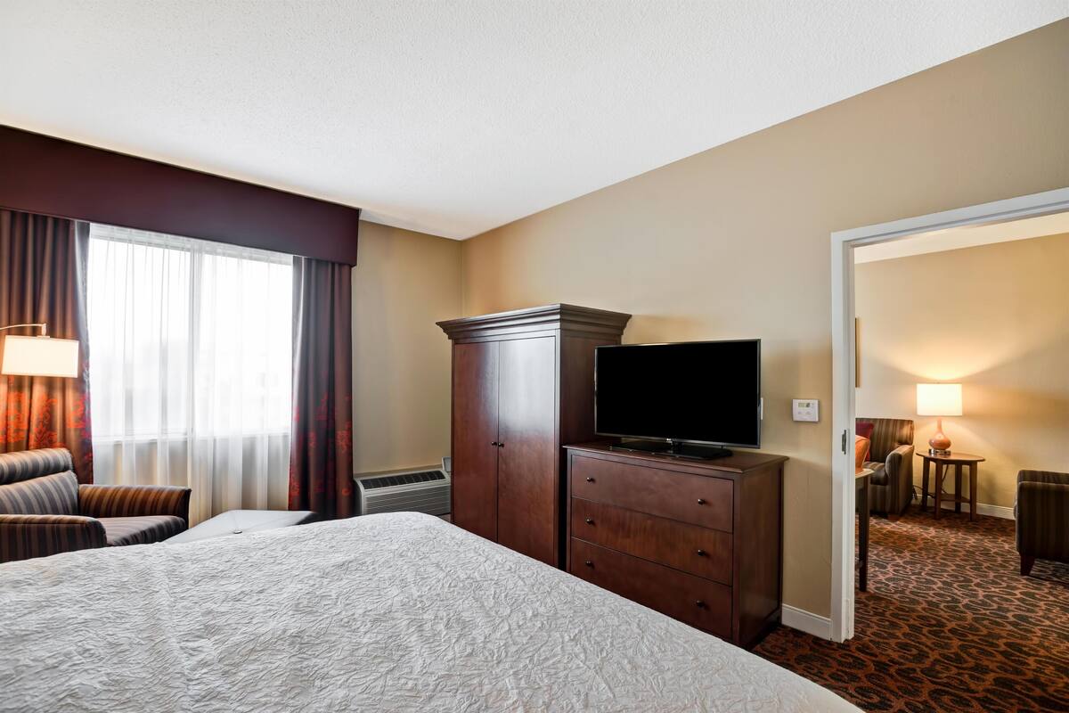 Una habitación de hotel con una cama grande, un televisor sobre un mueble de madera, un escritorio con una lámpara y vistas a otra habitación por una puerta abierta.