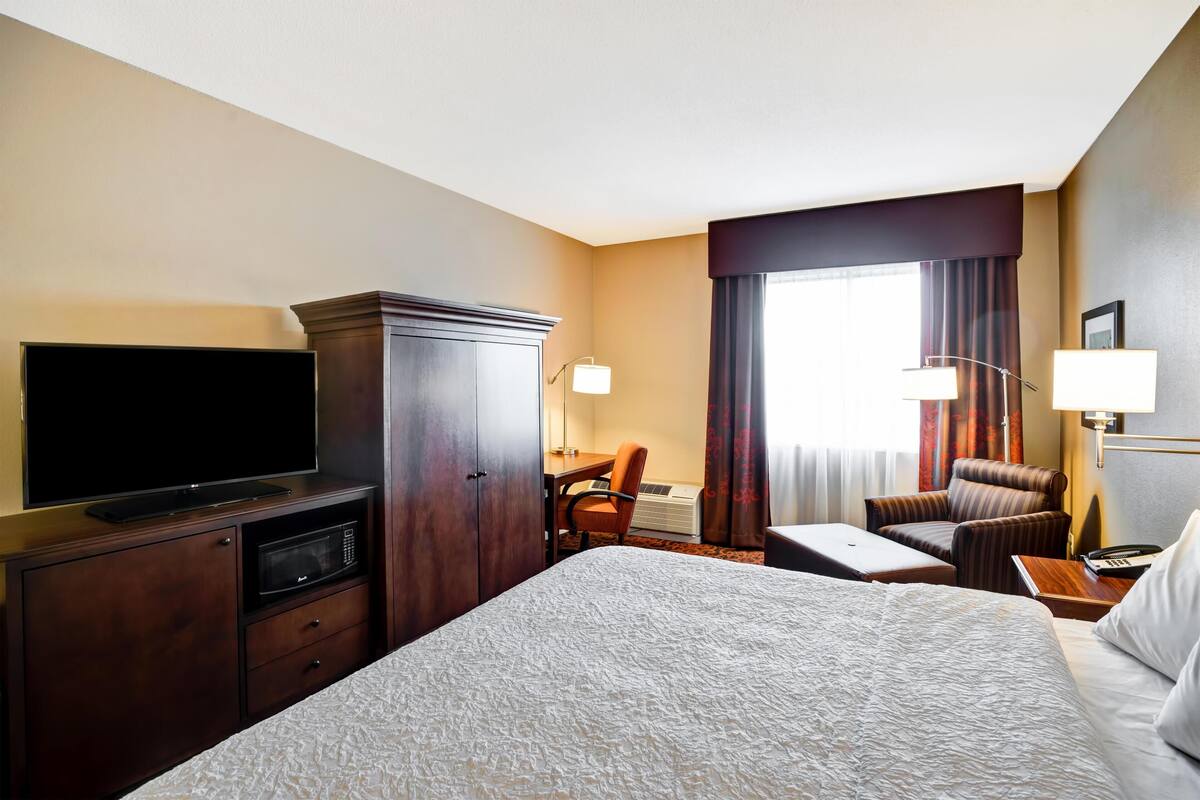 Habitación de hotel con una cama grande, un televisor sobre un mueble de madera, un escritorio con una silla y una ventana con cortinas.