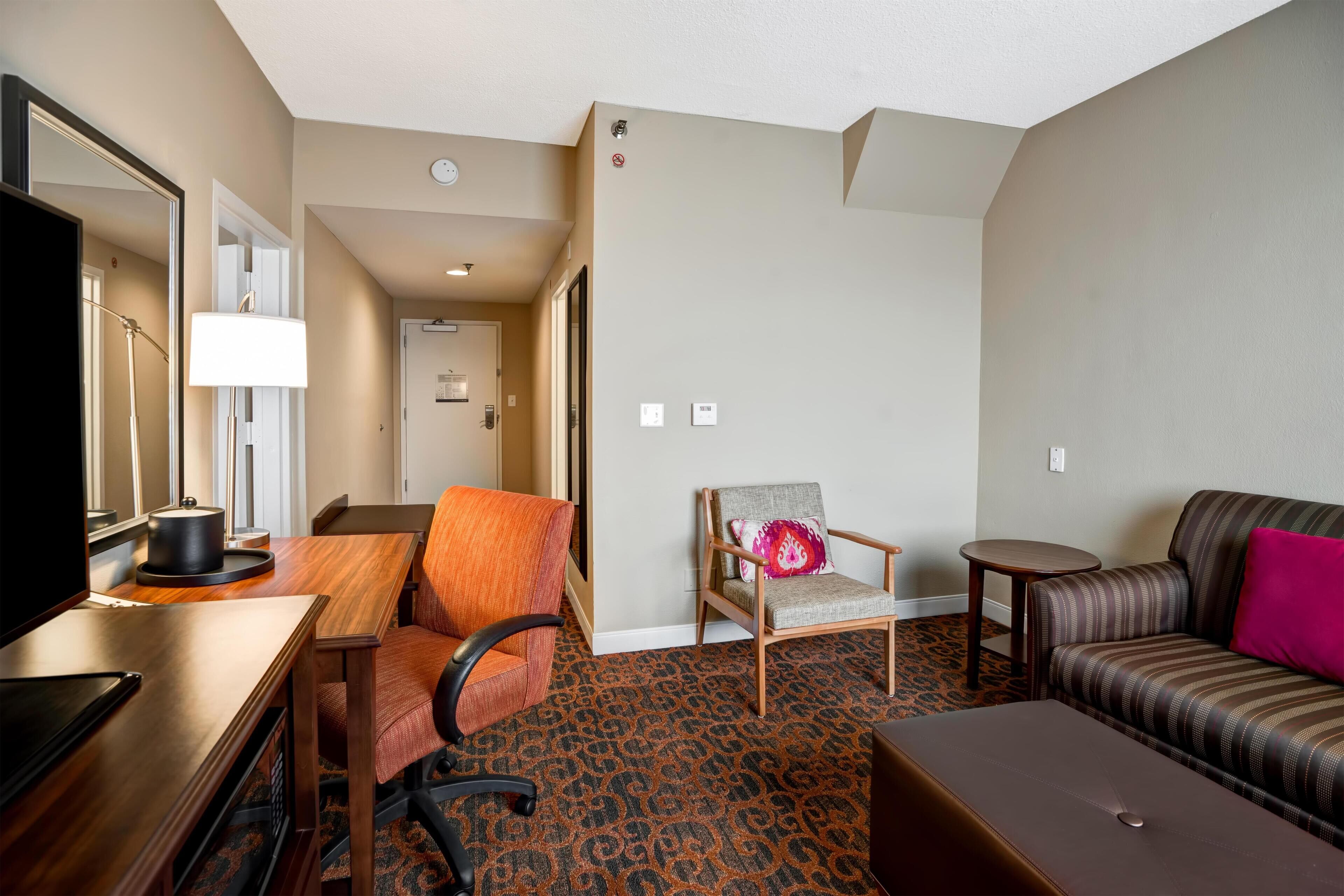 A hotel room with a wooden desk, a television, a sofa, a chair, and a patterned carpet.