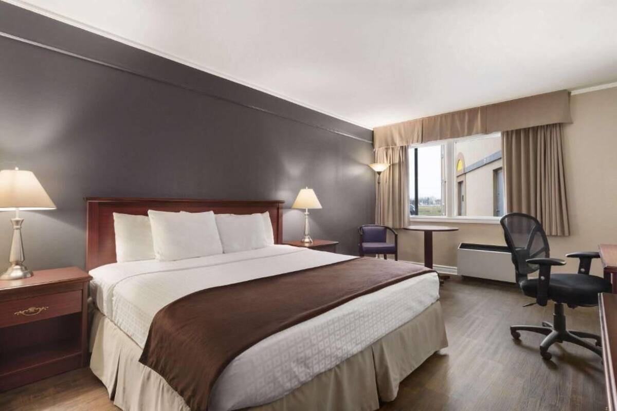 Ein Hotelzimmer mit einem großen Bett, zwei Nachttischen mit Lampen, einem Schreibtisch, einem Sessel und einem Fenster mit Vorhängen.