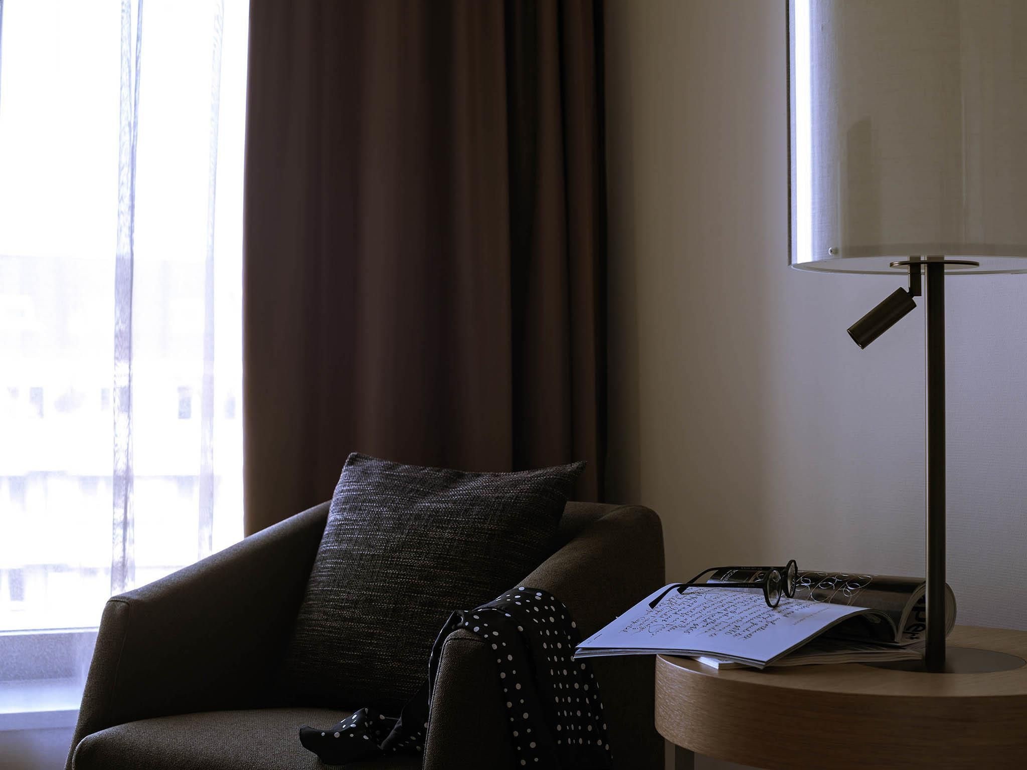Une chambre d’hôtel avec une fenêtre, une chaise, une lampe et une table de chevet sur laquelle se trouvent un livre et des lunettes.