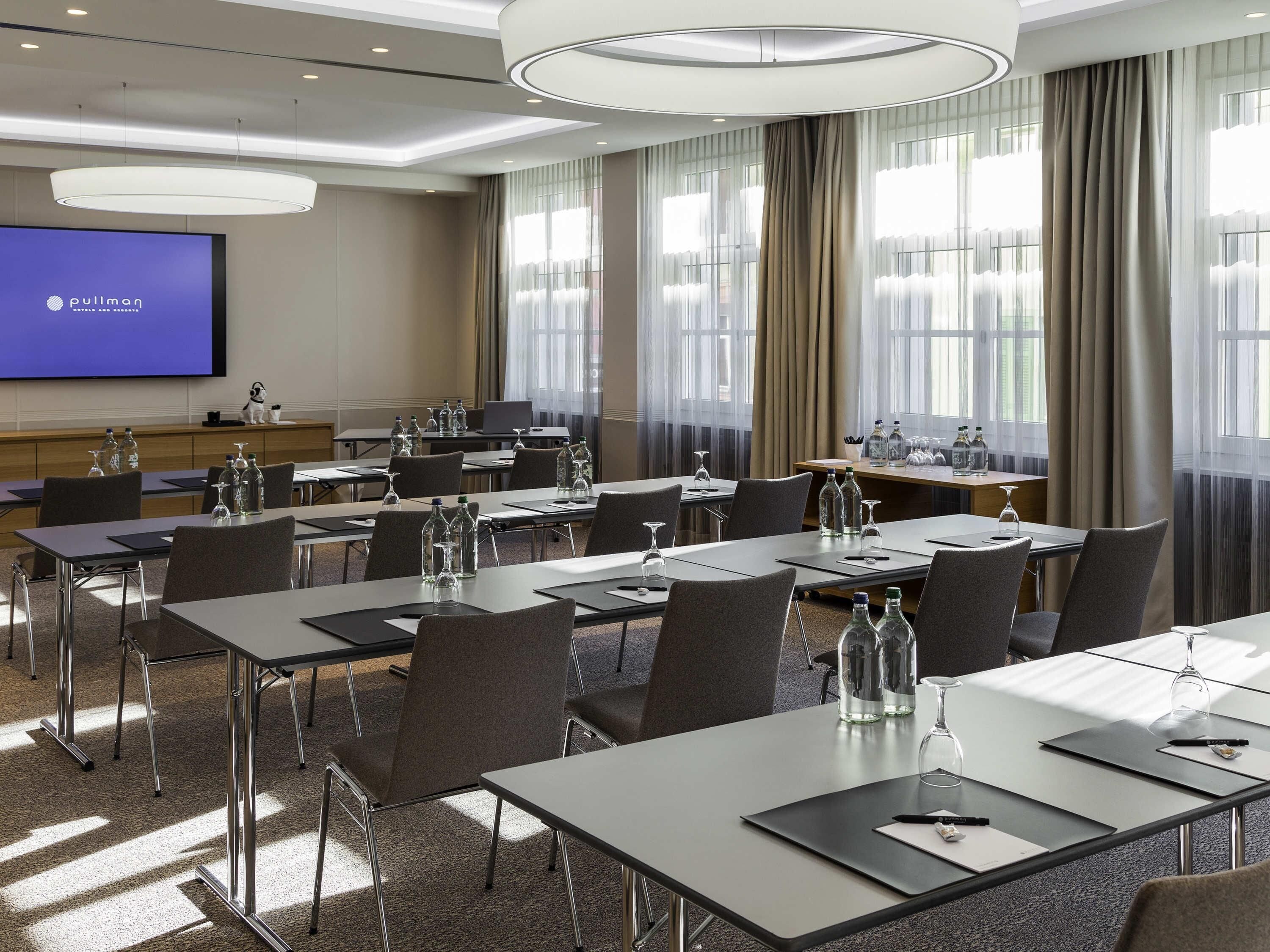 Een hotelconferentiezaal met tafels, stoelen, een groot scherm en uitzicht op het buitenlands gebied.