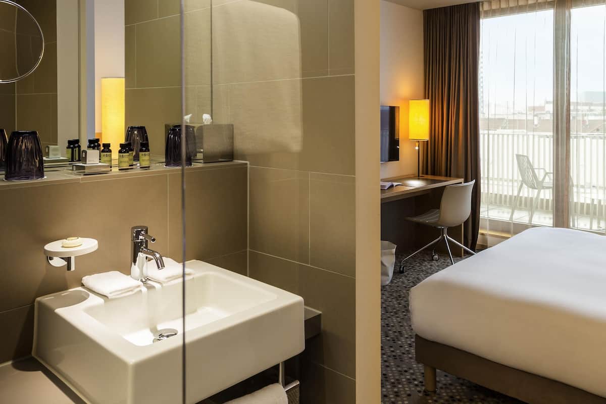 Ein Hotelzimmer mit einem großen Spiegel, einem weißen Waschbecken, einem Schreibtisch mit Stuhl, einem Bett und einem Balkon mit Blick.