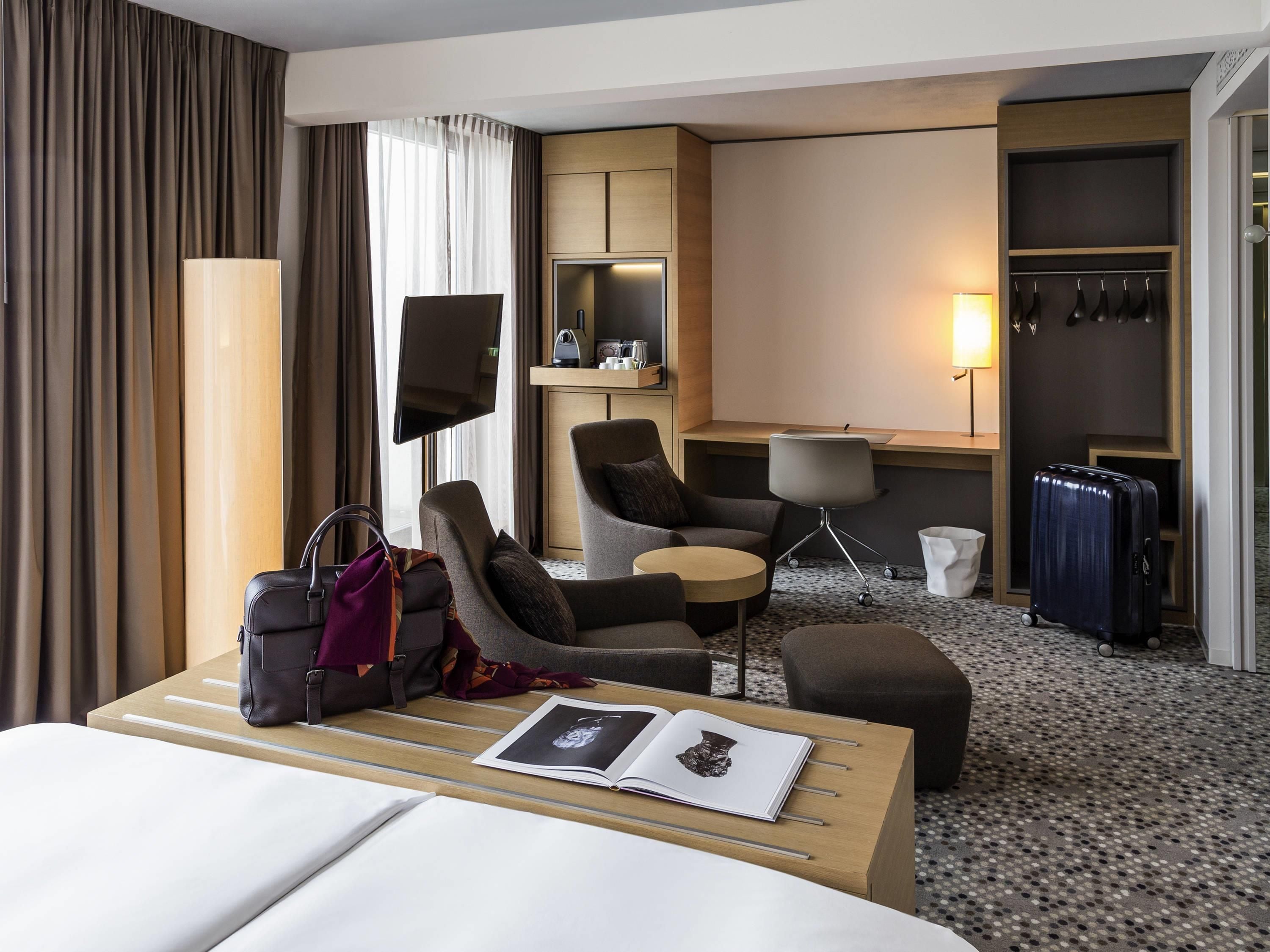A hotel room with a bed, a desk, a chair, a small table, a sofa, a TV, a wardrobe, and a bathroom.
