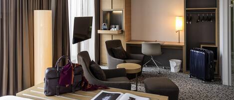 A hotel room with a bed, a desk, a chair, a small table, a sofa, a TV, a wardrobe, and a bathroom.