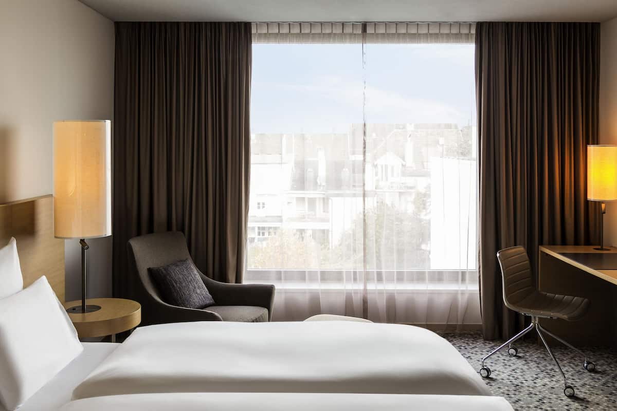 A hotel room with a bed, two chairs, a desk, and a large window with curtains.