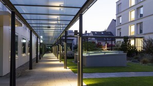 Une allée couverte avec des panneaux de verre, un chemin pavé et une zone herbeuse avec un banc.