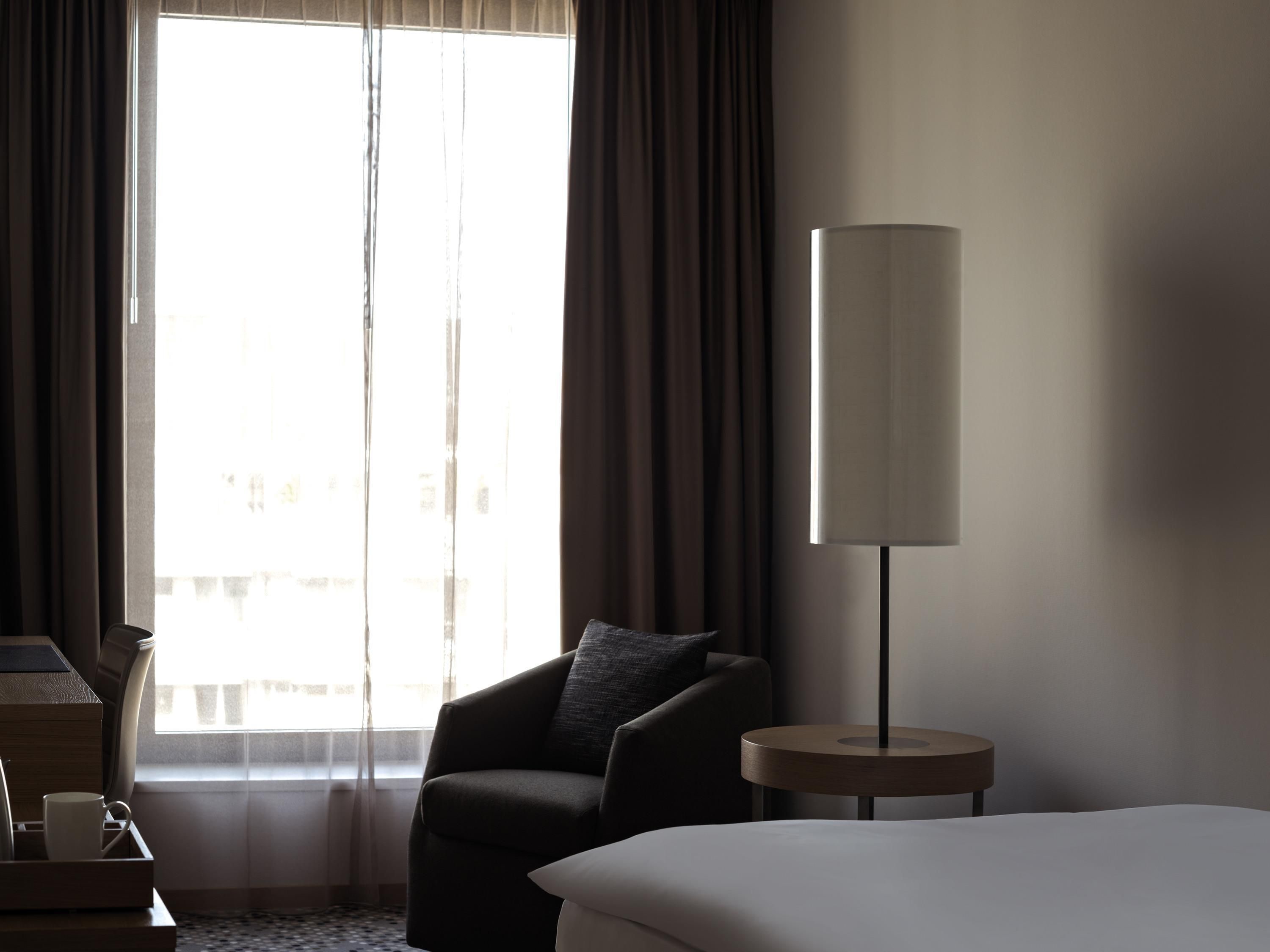 A hotel room with a large window, a chair, a bedside table, and a lamp.