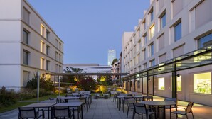 A modern outdoor patio area with multiple tables and chairs, surrounded by buildings with large windows and a glass structure overhead. - Pullman Basel Europe (Basel)