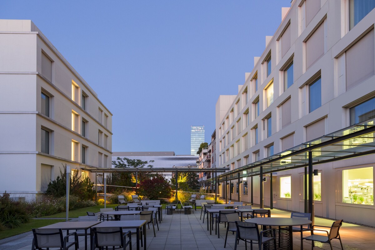 Un espace patio extérieur moderne, doté de plusieurs tables et chaises, entouré de bâtiments aux grandes fenêtres et surmonté d’une structure en verre.
