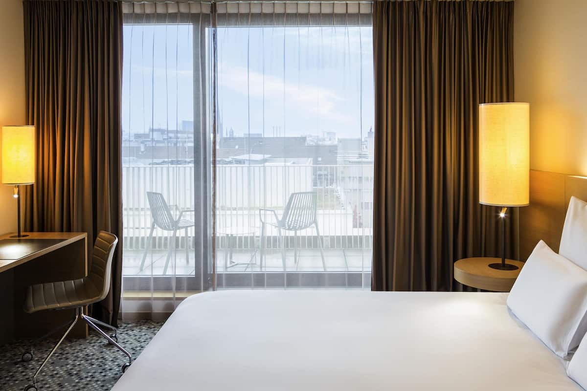 A hotel room with a large bed, two chairs, a desk, a lamp, and a view of the city through a large window.