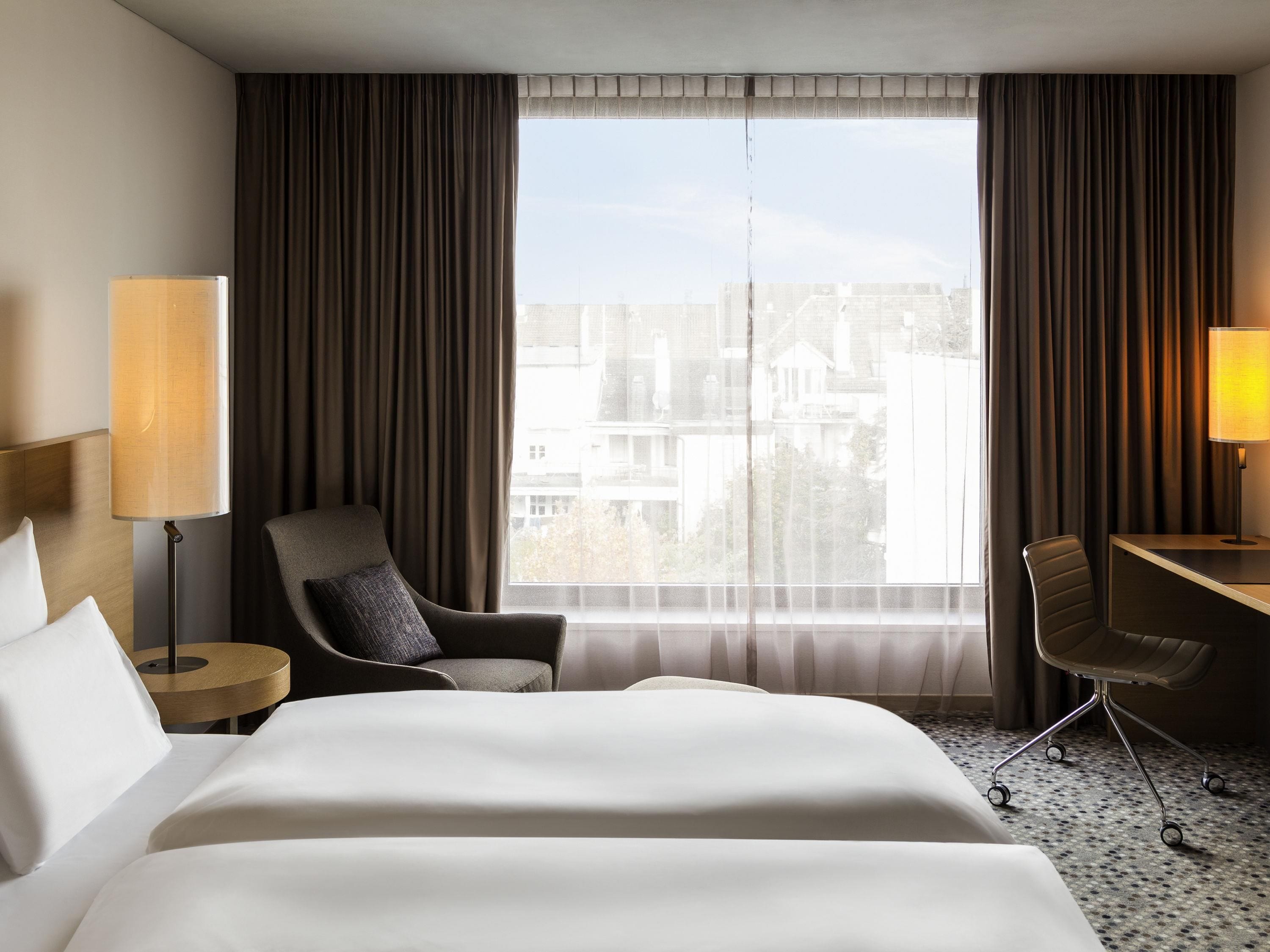 A hotel room with a bed, two chairs, a desk, and a large window with curtains.