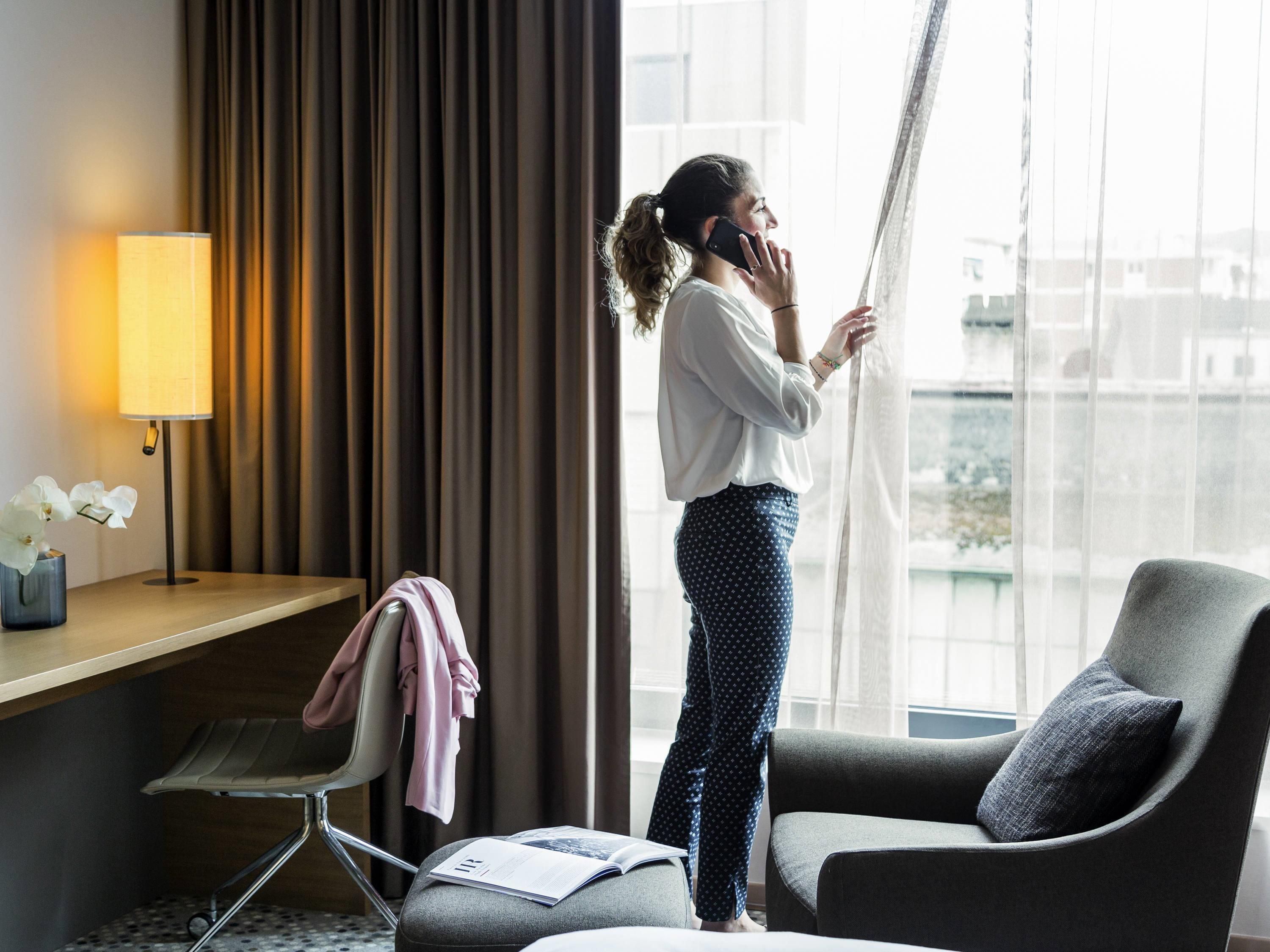 Ein Hotelzimmer mit großem Fenster, Schreibtisch mit Lampe, Stuhl und Bett.