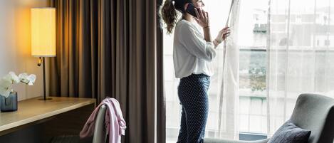 A hotel room with a large window, a desk with a lamp, a chair, and a bed.