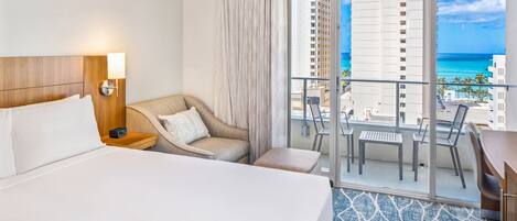 A hotel room with a large bed, a sofa, a small table, and a balcony with a view of the ocean and buildings.