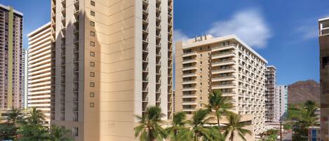 A high-rise hotel with multiple balconies, a central courtyard with palm trees, and a view of a mountain in the background.