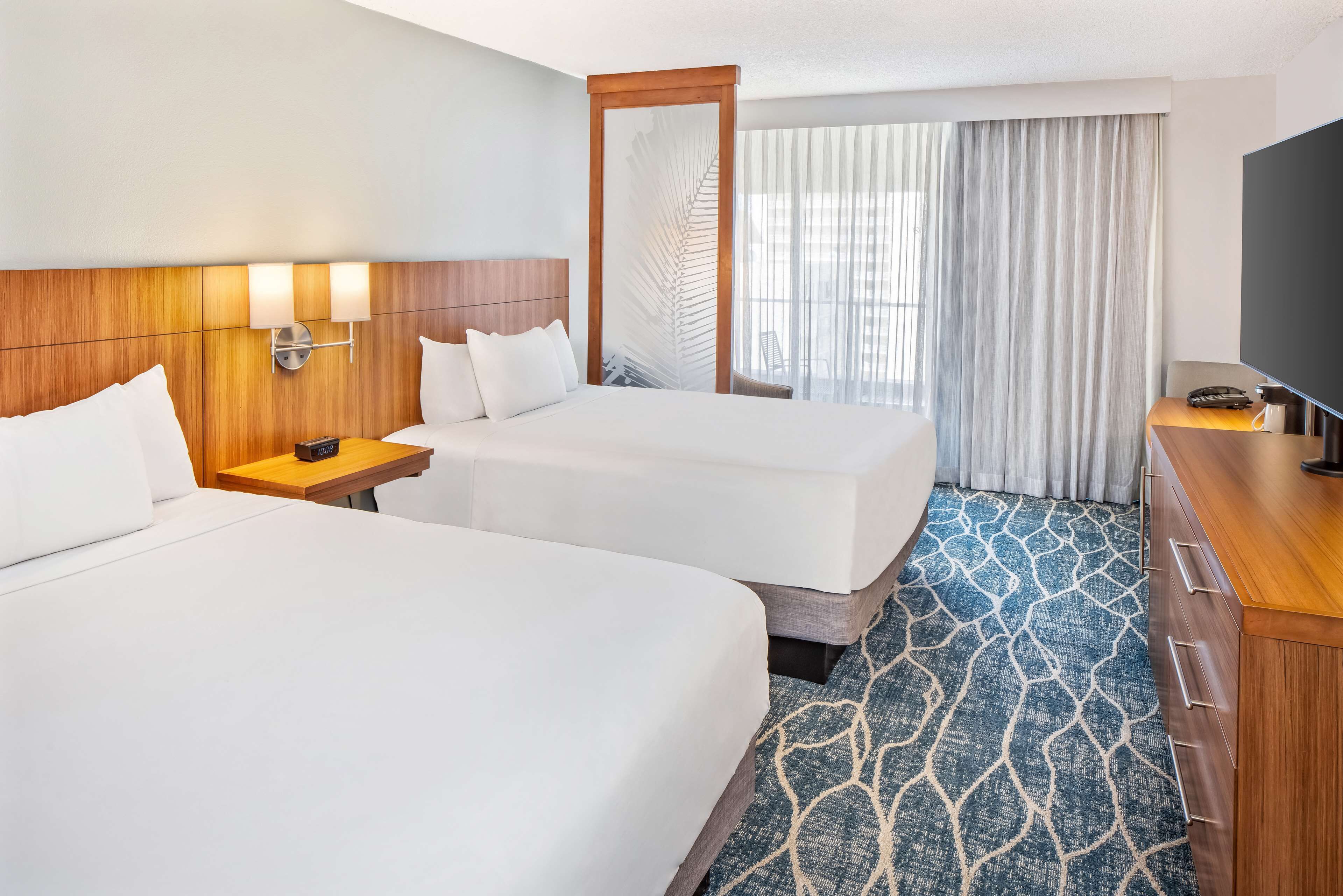 A hotel room with two beds, a wooden headboard, a nightstand, a TV, and a large window with curtains.