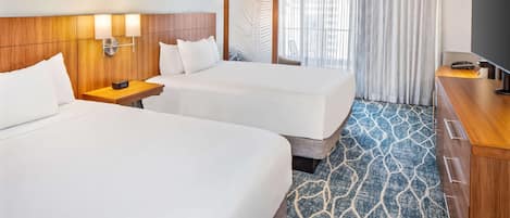 A hotel room with two beds, a wooden headboard, a nightstand, a TV, and a large window with curtains.