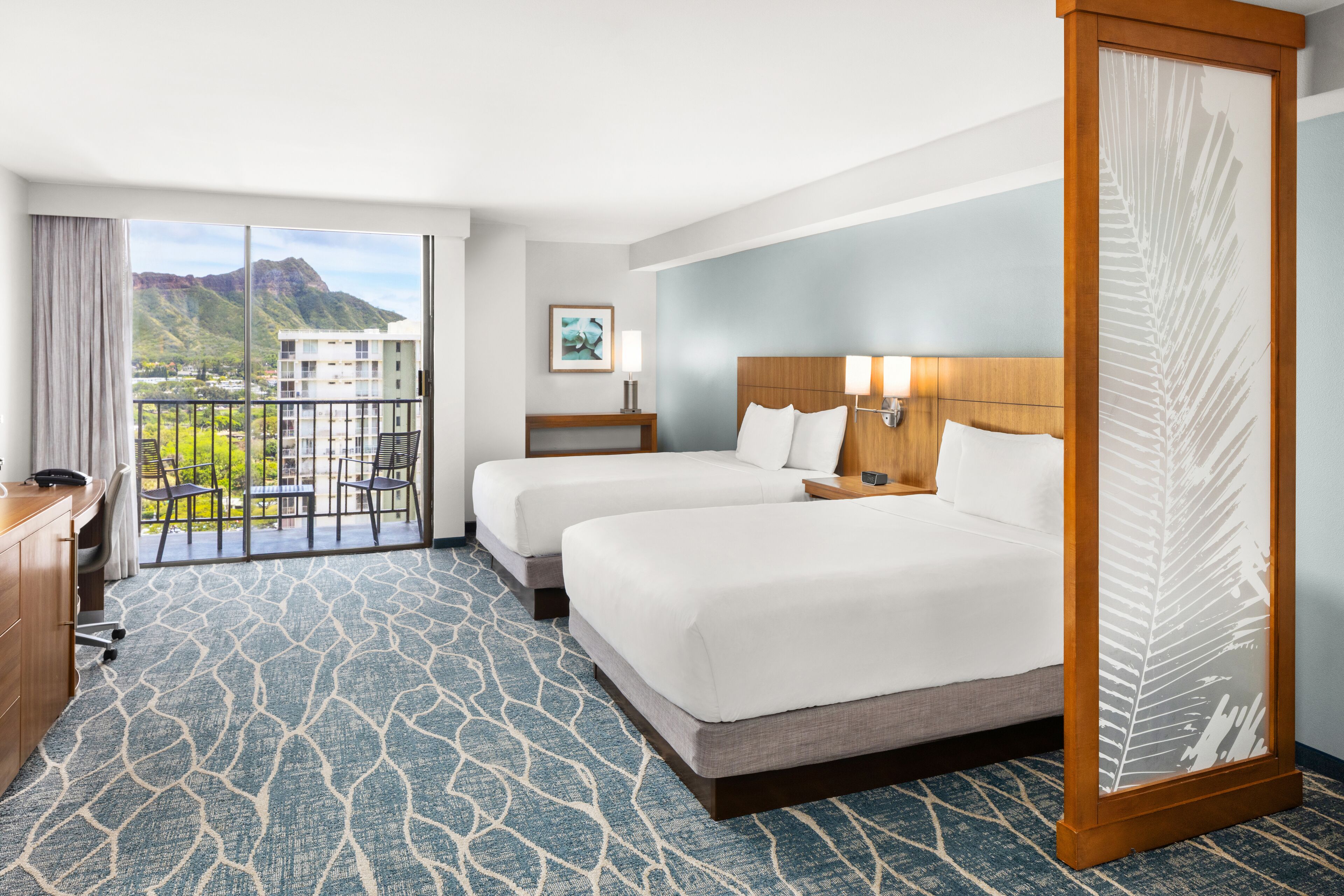 A hotel room with two beds, a desk, a chair, a large window with a view of mountains, and a balcony.