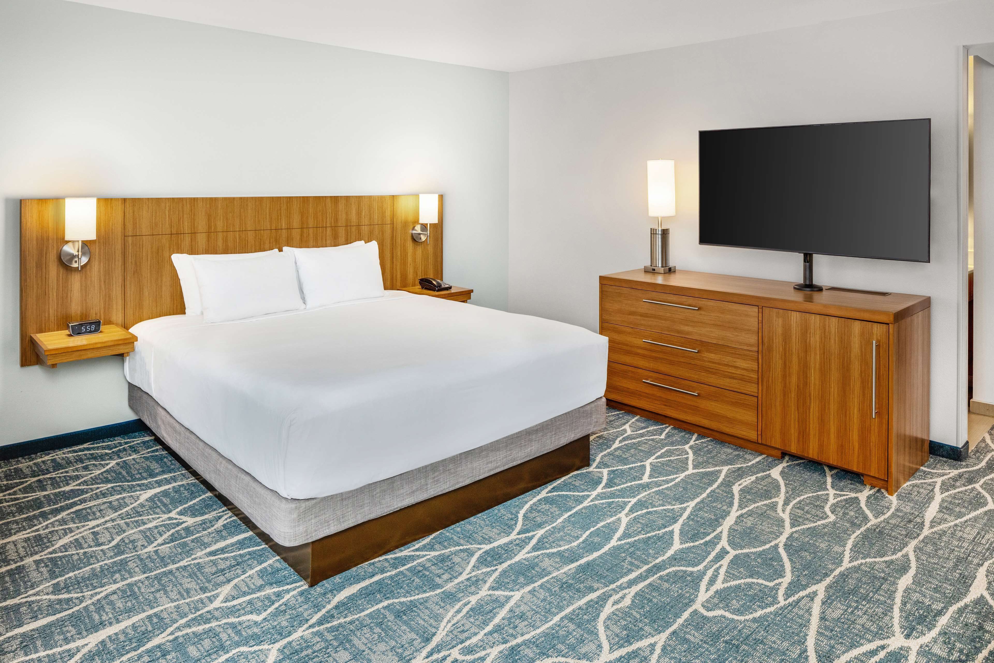 A hotel room with a large bed, wooden headboard, two bedside tables with lamps, a flat-screen TV on a wooden cabinet, and a patterned carpet.