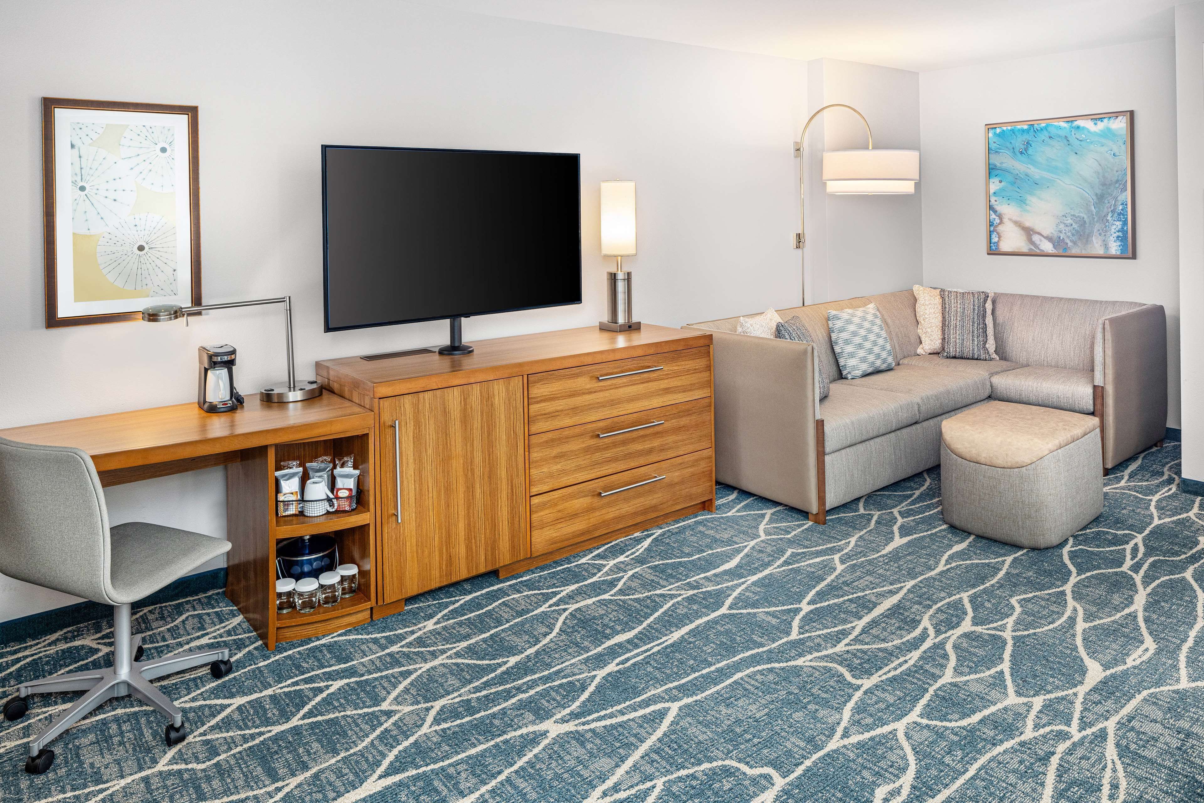 Ein Hotelzimmer mit Flachbildfernseher, Holztisch, Stuhl und einer Couch mit passendem Hocker.