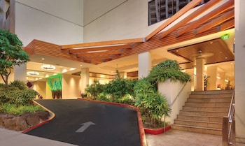 A bar area with wooden stools, a counter, and a television screen. There are chairs and tables in the foreground. at Hyatt Place Waikiki Beach
