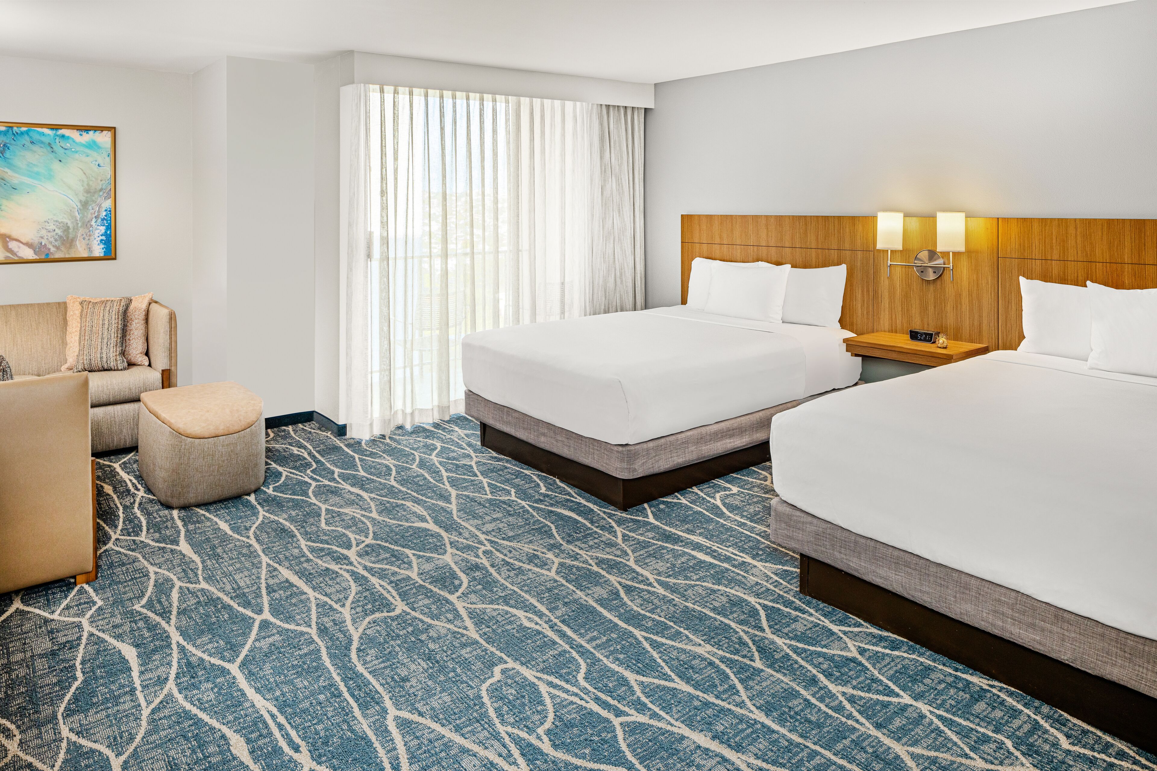 Quarto de hotel com duas camas, um sofá, um pequeno banquinho, uma mesa de cabeceira com relógio e uma janela grande com cortinas leves.