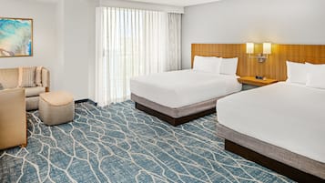 Quarto de hotel com duas camas, um sofá, um pequeno banquinho, uma mesa de cabeceira com relógio e uma janela grande com cortinas leves.