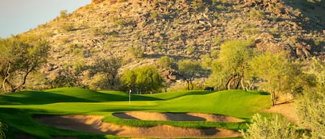 Un campo de golf con búnkeres de arena, árboles y una colina rocosa al fondo.