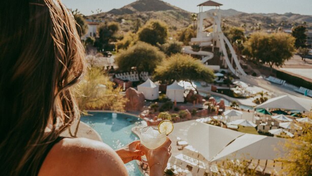 A person holding a drink near a pool with a water slide and outdoor seating in the background.