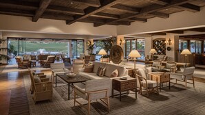 A spacious living area with a high ceiling and wooden beams, furnished with wicker chairs, sofas, and tables. Large windows offer a view of a golf course.