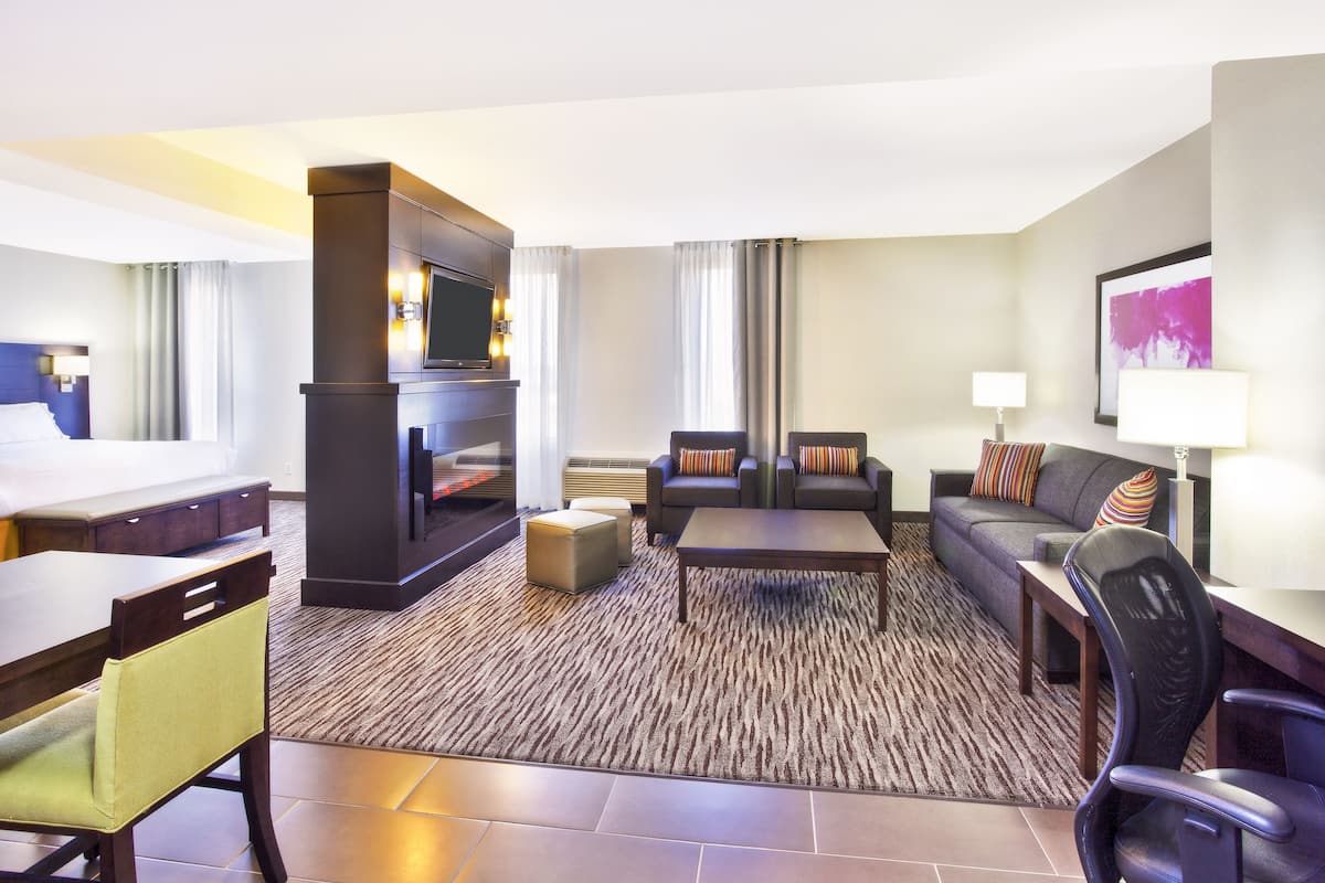 A hotel room with a living area featuring a sofa, armchairs, a coffee table, a flat-screen TV, a fireplace, and a dining table with chairs.