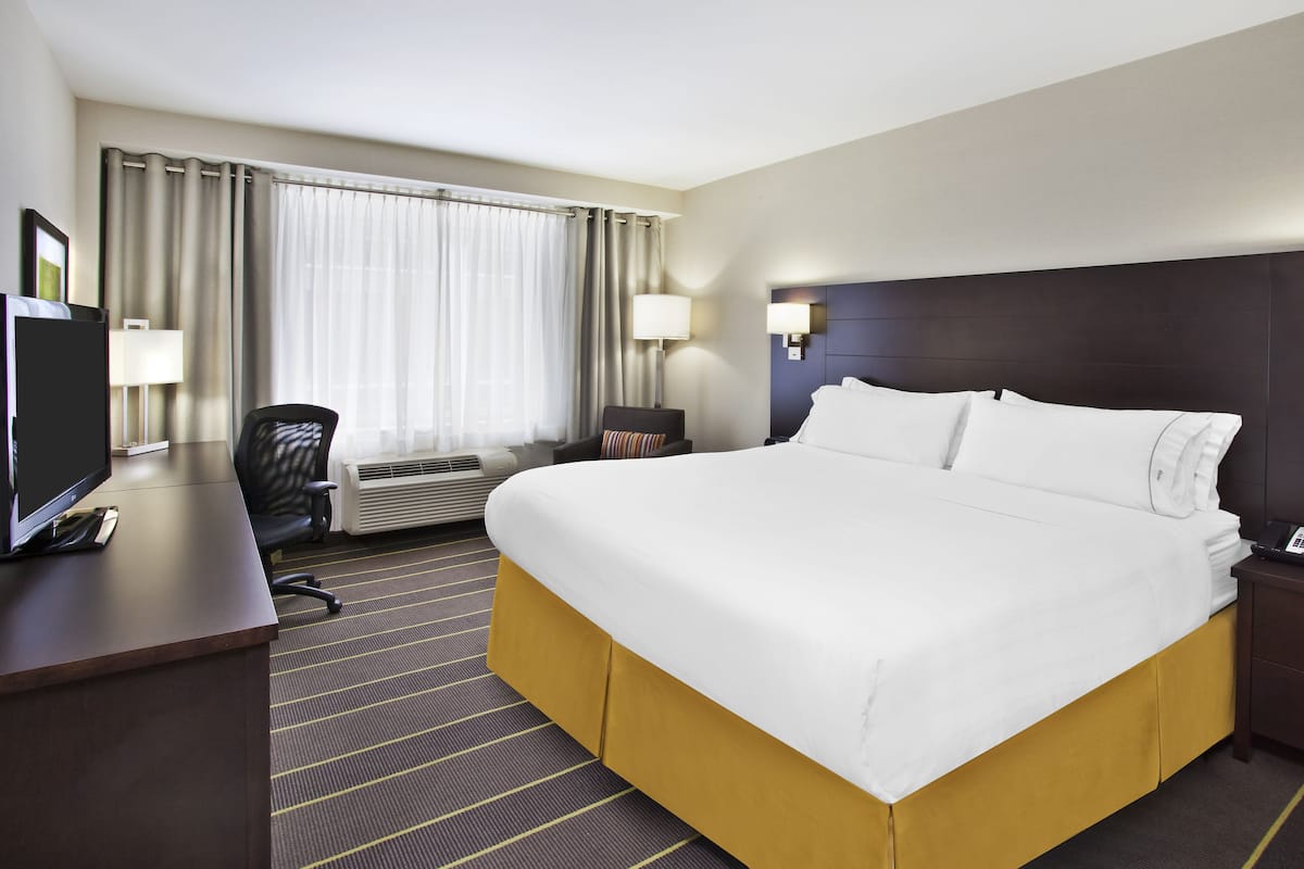 A hotel room with a large bed, a desk with a computer, a chair, a lamp, and a window with curtains.