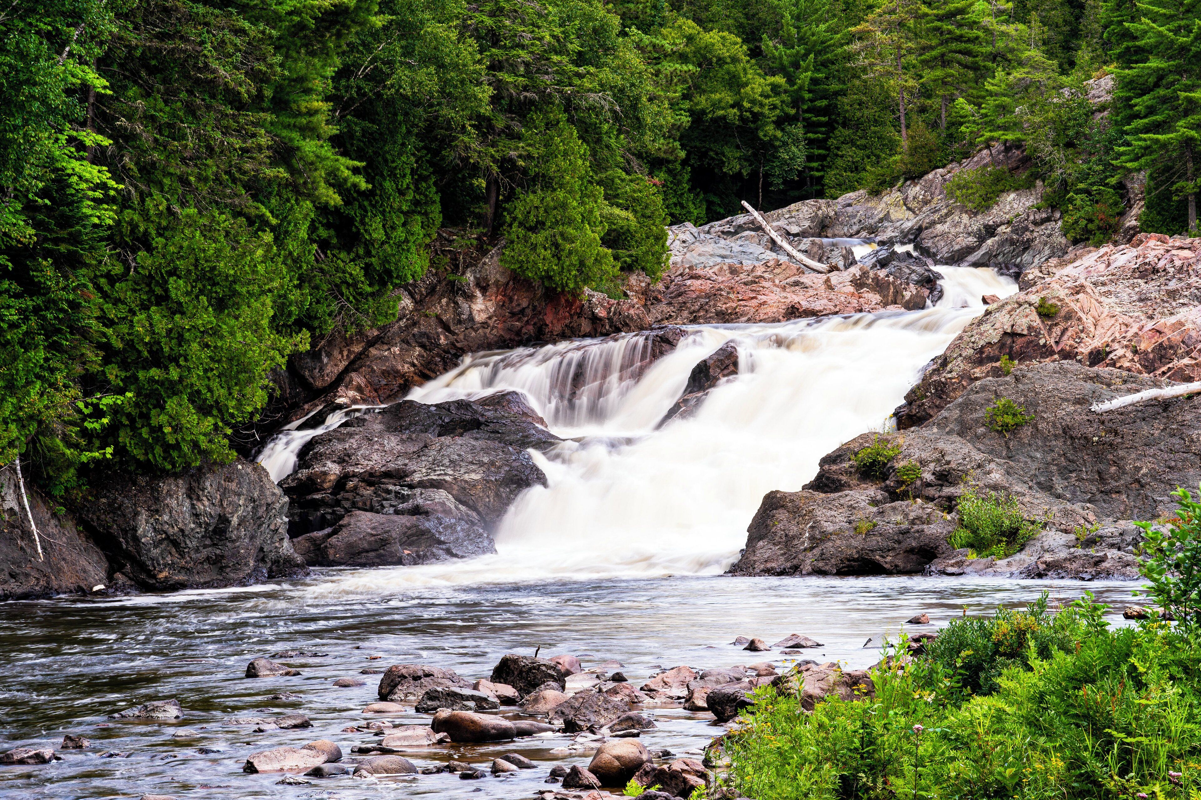A waterfall cascading over rocks into a river, surrounded by dense green forest.