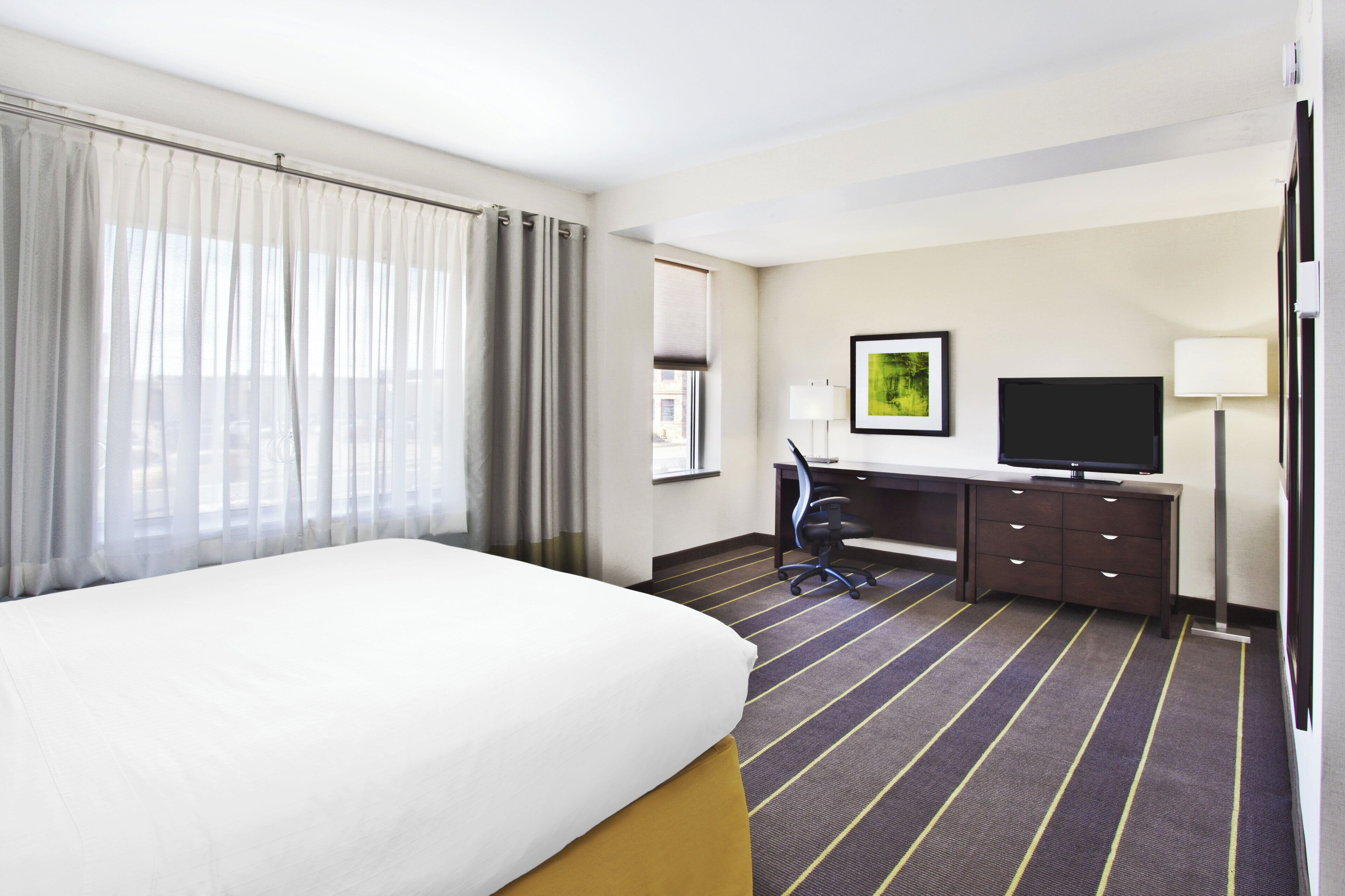 Habitación de hotel con cama, escritorio, silla, televisor y ventana con cortinas.
