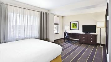 Habitación de hotel con cama, escritorio, silla, televisor y ventana con cortinas.