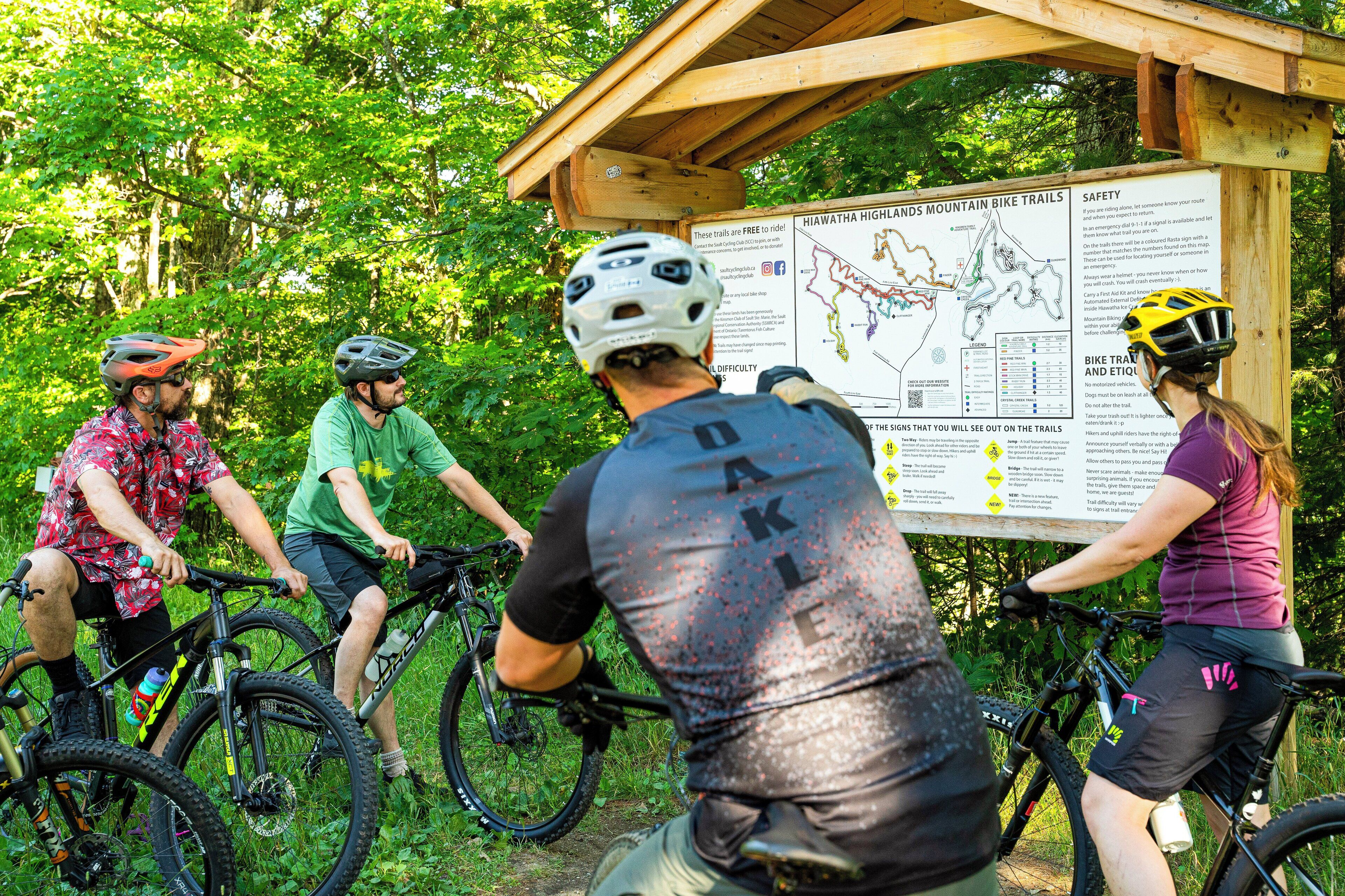 A group of people on mountain bikes at a trailhead with a map and safety information board.