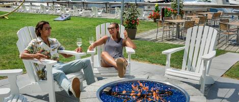 Two people seated at a table with a fire pit in the center, surrounded by white Adirondack chairs, with a marina and boats in the background.