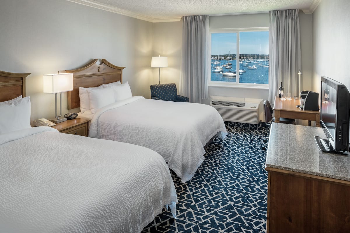 A hotel room with two beds, a desk, a TV, a lamp, and a view of the marina.